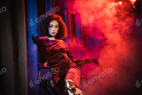Preview: african american woman in velvet dress and gloves looking at camera while sitting on chair
