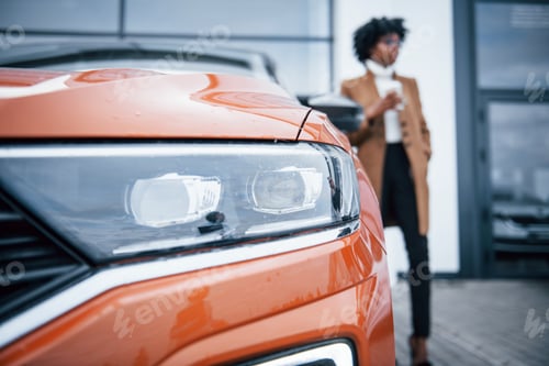 Visualização: Com uma xícara de bebida. Jovem afro-americana em óculos fica ao ar livre perto de um carro moderno