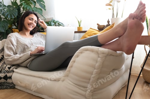 Preview: Happy woman using laptop lying on the sofa at home cozy living room.