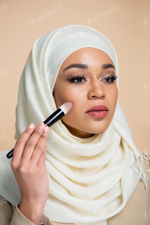 Preview: young muslim woman in hijab applying face foundation isolated on beige