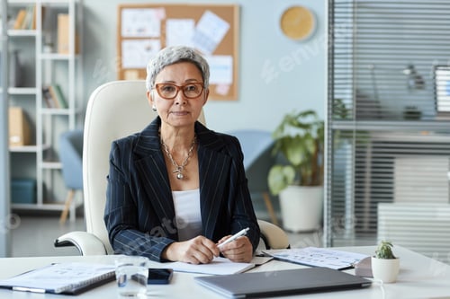 Preview: Senior woman as female boss sitting at workplace in office and looking at camera