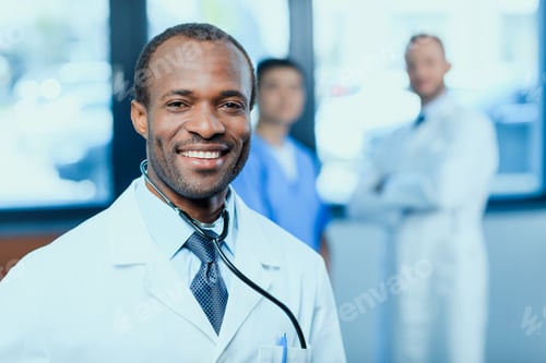 Preview: portrait of smiling doctor with stethoscope with colleagues behind in clinic