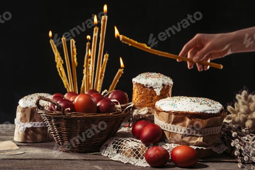 Preview: cropped shot of person holding burning candle and easter cakes with painted eggs on table