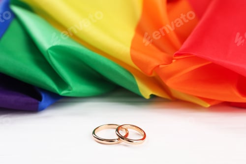 Preview: Rainbow LGBT flag and wedding rings on white wooden table, closeup