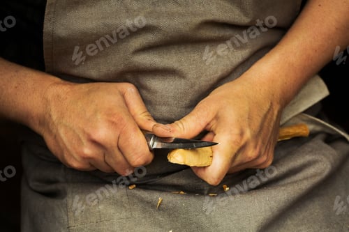 Preview: Close up of a craftsman's hands pressing and shaping a small piece of wood into a spoon with a sharp