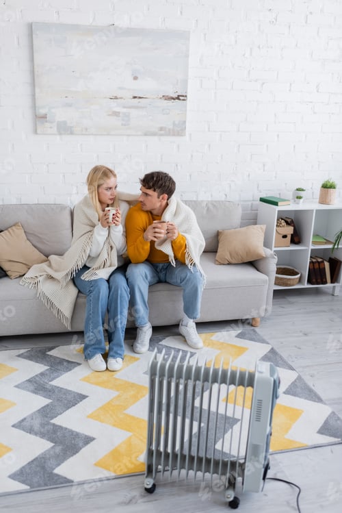 Preview: young couple covered in blanket sitting with cups of tea near radiator heater
