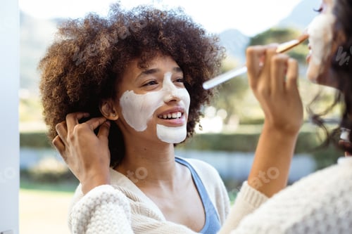 Preview: Smiling biracial female friends applying beauty cream on each others faces in balcony