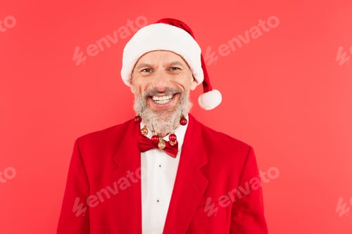 Preview: happy mature man in santa hat with christmas balls on beard isolated on red