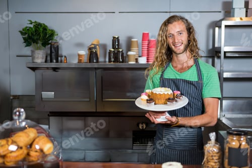 Preview: Man smiling, holding bundt cake on white stand wearing striped apron behind bakery counter