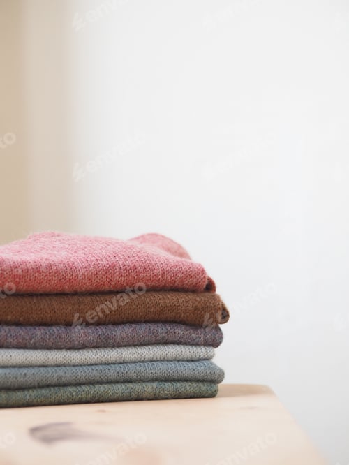 Preview: A stack of multi-colored knitted clothes lies on a wooden table in a bright room.