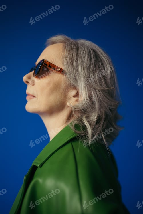 Preview: profile of senior grey haired lady in trendy sunglasses and green leather jacket isolated on blue