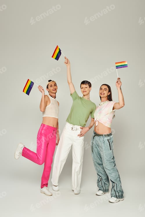 Preview: happy and young lgbtq friends with tattoos standing in colorful clothes and holding rainbow flags