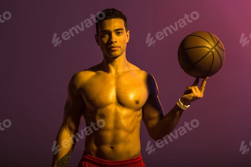 Preview: good-looking sportive mixed race man holding brown ball and looking at camera on purple background