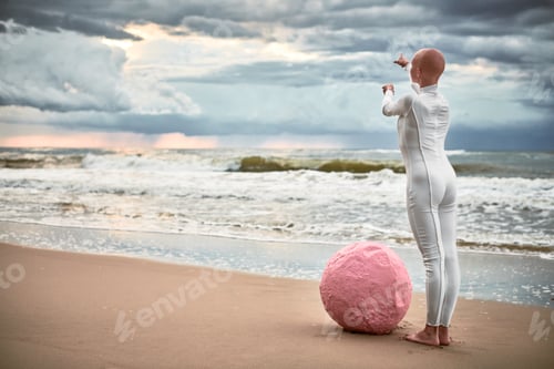 Preview: Hairless girl with alopecia in white futuristic suit standing on stone sea beach stretched out arms