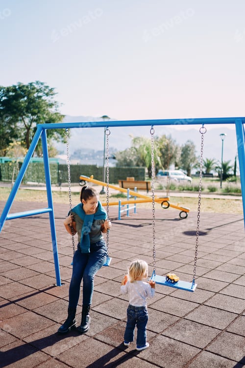 Preview: Mom sits on a chain swing in the playground and looks at a little girl standing next to her