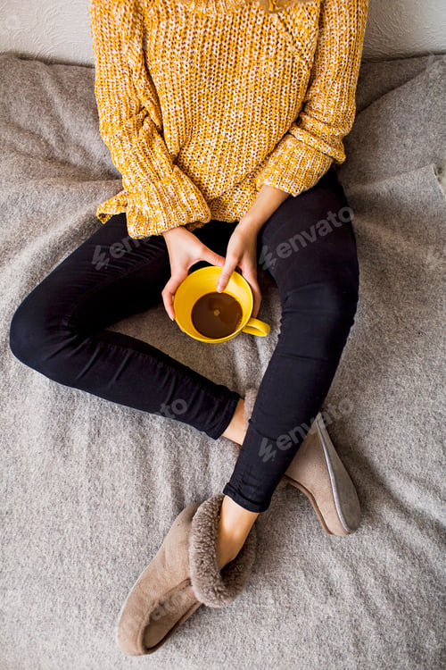 Preview: Low section of woman holding coffee cup while resting on bed