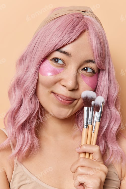 Preview: Pretty thoughtful Asian woman with long pink dyed hair holds cosmetic brushes looks away applies col