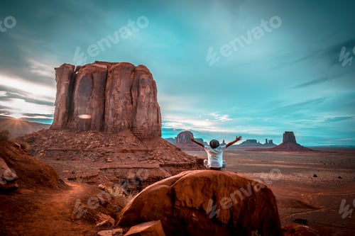 Preview: A boy in a white T-shirt sitting on a stone with his arms up in Monument Valley