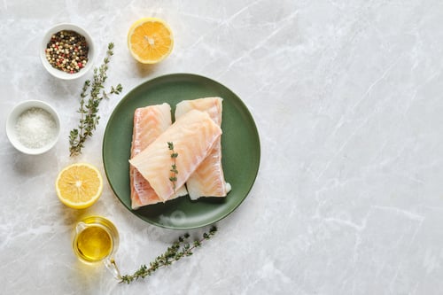 Preview: Overhead view of fresh cod fish fillets on a plate with herbs and citrus on a marble countertop