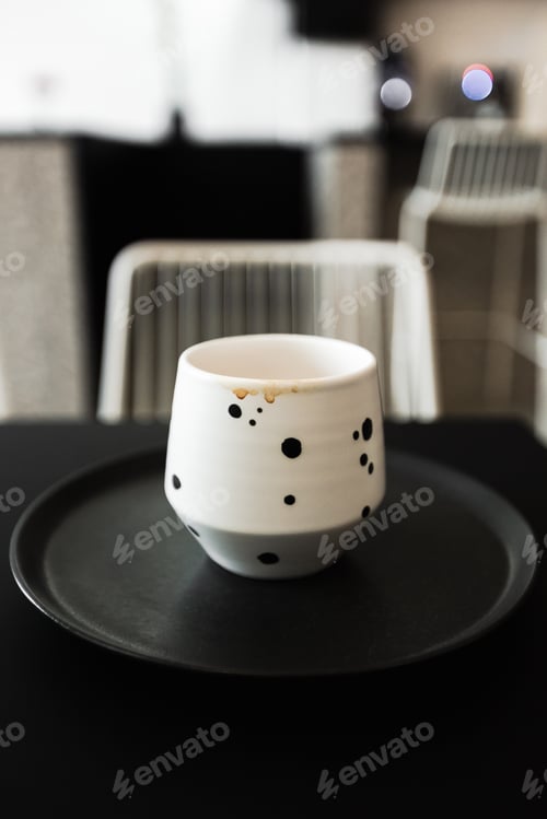 Preview: A white cup with drops of coffee on it, on a black plate, on a black table, in a cafe