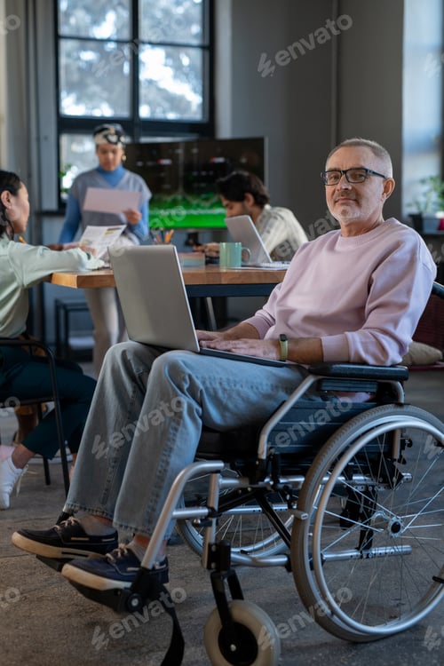 Preview: Mature employee with laptop on his knees sitting in wheelchair