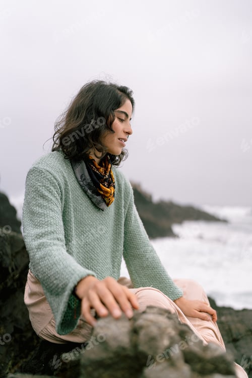 Preview: Young woman wearing wool sweater sitting on rocks at the beach in winter
