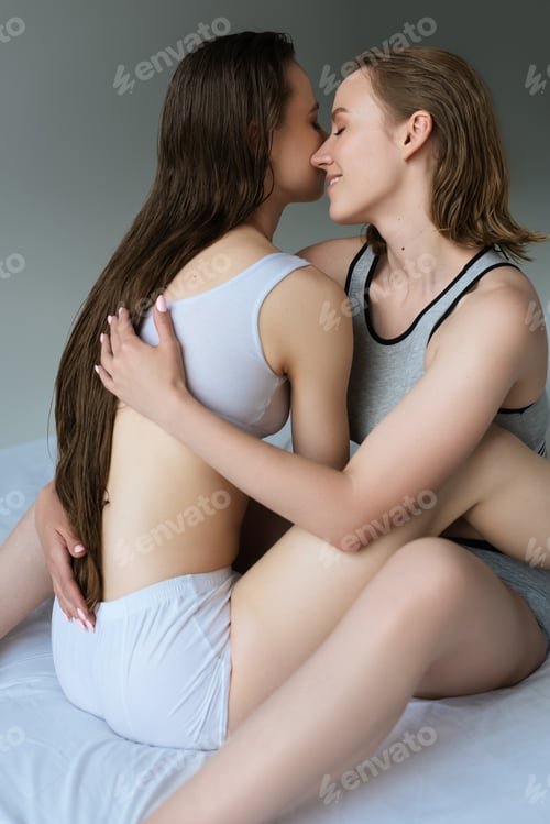 Preview: smiling lesbian woman embracing long haired girlfriend while sitting on bed isolated on grey