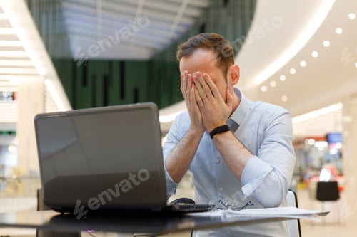 Preview: Feeling tired. Frustrated young man covering his face with hands while working on laptop