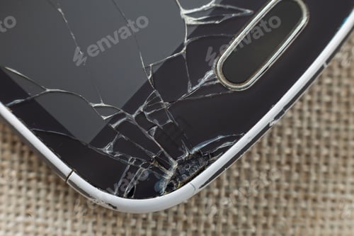 Preview: Close-up detail of black old cellphone with cracked screen on light cloth background.