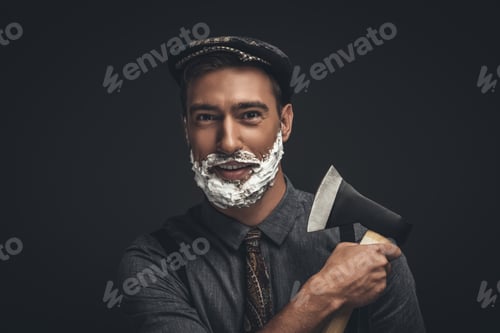 Preview: Smiling young man in flat cap and with shaving cream on his face, grooming his beard with an axe