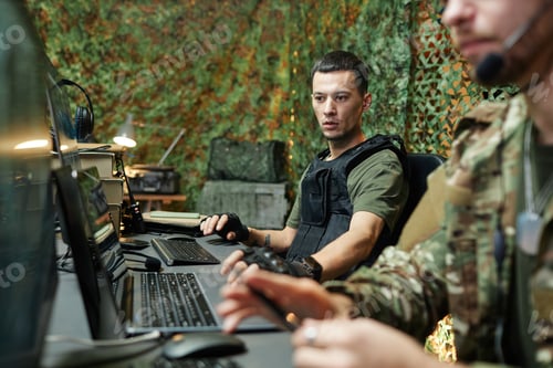 Preview: Young serious male military officer in body armor sitting by workplace