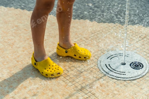 Preview: Children enjoying summer pool with water jets, summer vacation. Yellow child's flip flops