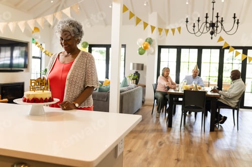 Preview: Elderly woman carrying birthday cake in living room while family enjoys celebration