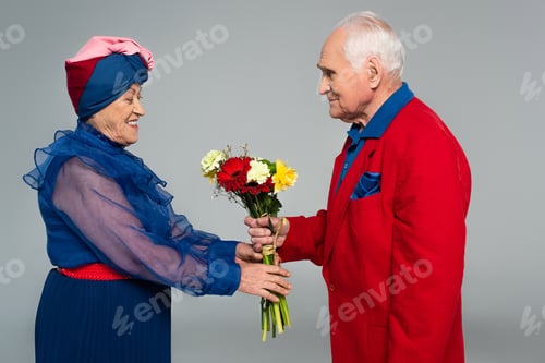Preview: smiling elderly man in red blazer presenting bouquet of flowers to wife in blue dress and turban