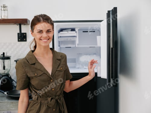 Preview: Woman smiling with teeth looking into camera in kitchen at home opened freezer empty with ice inside