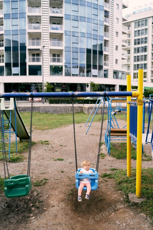 Preview: Little girl sits on a swing on a playground in the courtyard of an apartment building