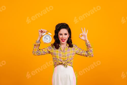 Preview: Pretty young woman posing with clock and okay sign. Smiling pinup girl in checkered shirt standing