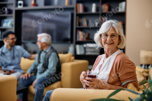 Preview: Happy senior woman drinking tea at residential care home and looking at camera.
