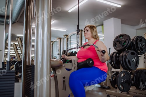 Preview: Mid adult plus size woman working out on a lat pulldown machine indoors in gym