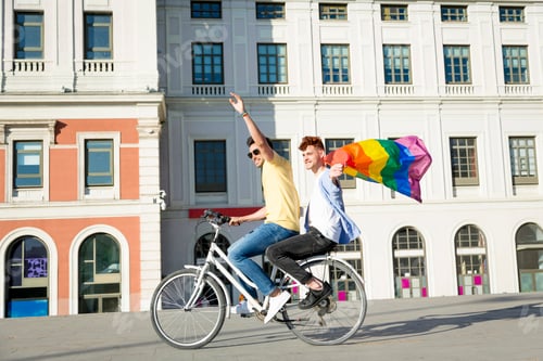 Preview: Young gay men couple having fun riding bicycles with gay pride flag outdoors. lgbt concept