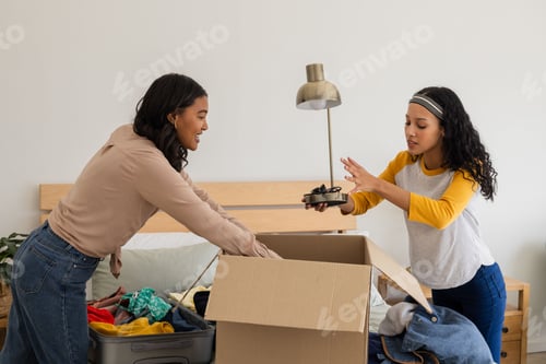 Preview: Mother and daughter packing clothes into cardboard box at home, smiling together