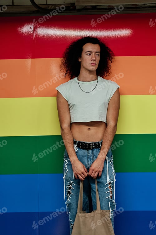 Preview: Fashionable gay man leaning against a wall in the colours of the gay pride flag