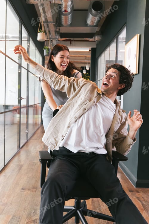 Preview: Young woman having fun pushing man on chair along walkway in open plan office