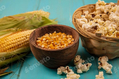 Preview: top view of corn kernels on a wooden bowl with fresh corns with popcorns on a bucket on a blue