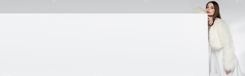 Preview: pretty young woman in white faux fur jacket leaning on cube while posing on grey, banner