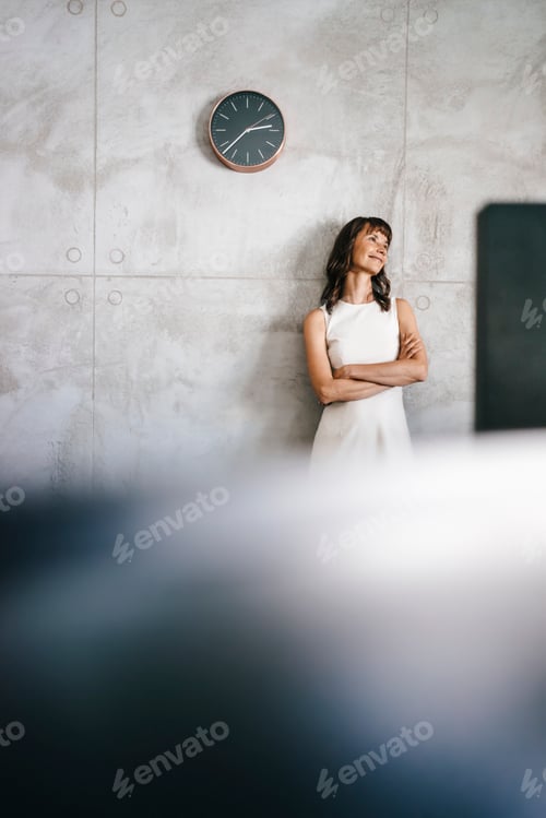 Preview: Businesswoman standing under wall clock with arms crossed