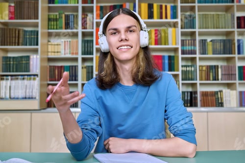 Preview: Teenage college student in headphones looking at webcam, sitting at desk in library