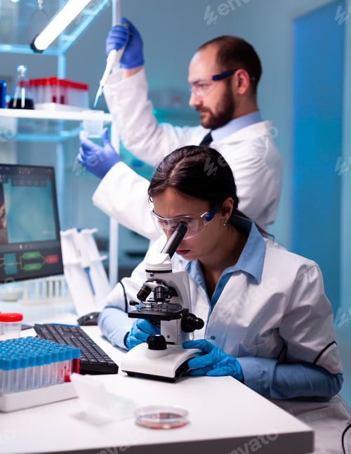 Preview: Scientist looking at blood sample through microscop and assitant taking liquid