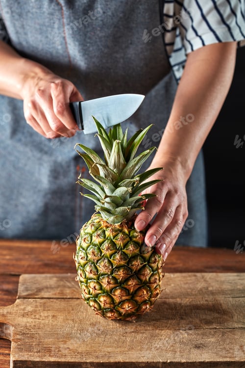 Preview: Woman's hands with a knife cut a juicy pineapple on a wooden board on a brown table around a