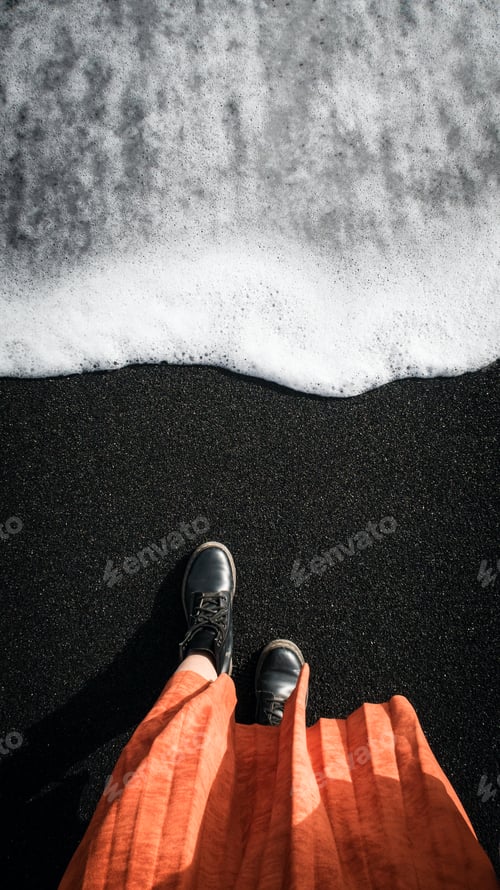 Preview: Woman in orange coat at the black volcanic sand.